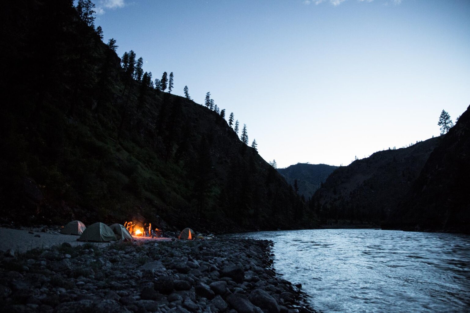 Twilight on the river with camp set up down the beach and a group of people standing around a campfire.