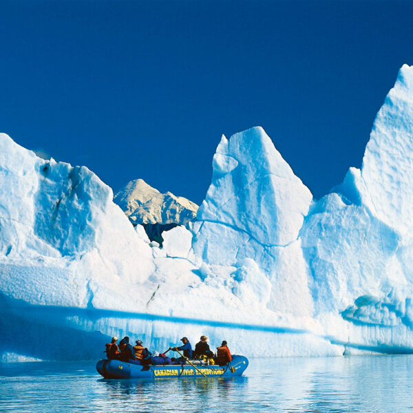 A raft full of people floats in front of massive glaciers on the Alsek River.