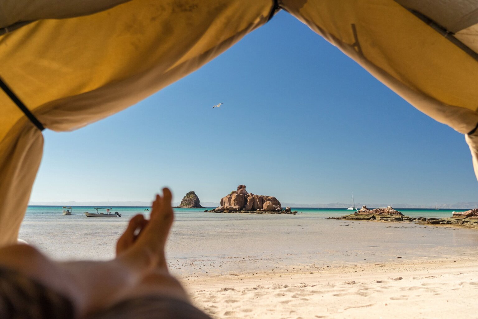 A serene beach view from inside a tent, showcasing turquoise waters, sandy shores, and rock formations under a clear blue sky.