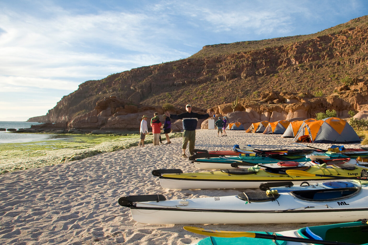 A line of sea kayaks and tents along a beach as guests mingle on the sand at camp.