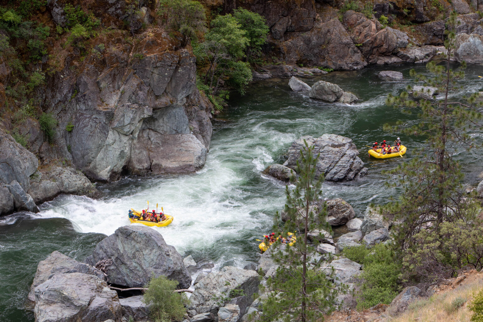 A yellow raft full of paddlers makes their way through a big rapid as two other boats watch from downstream.