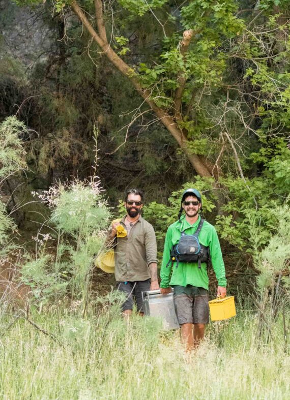 two men carrying a portable toilet out of the woods.