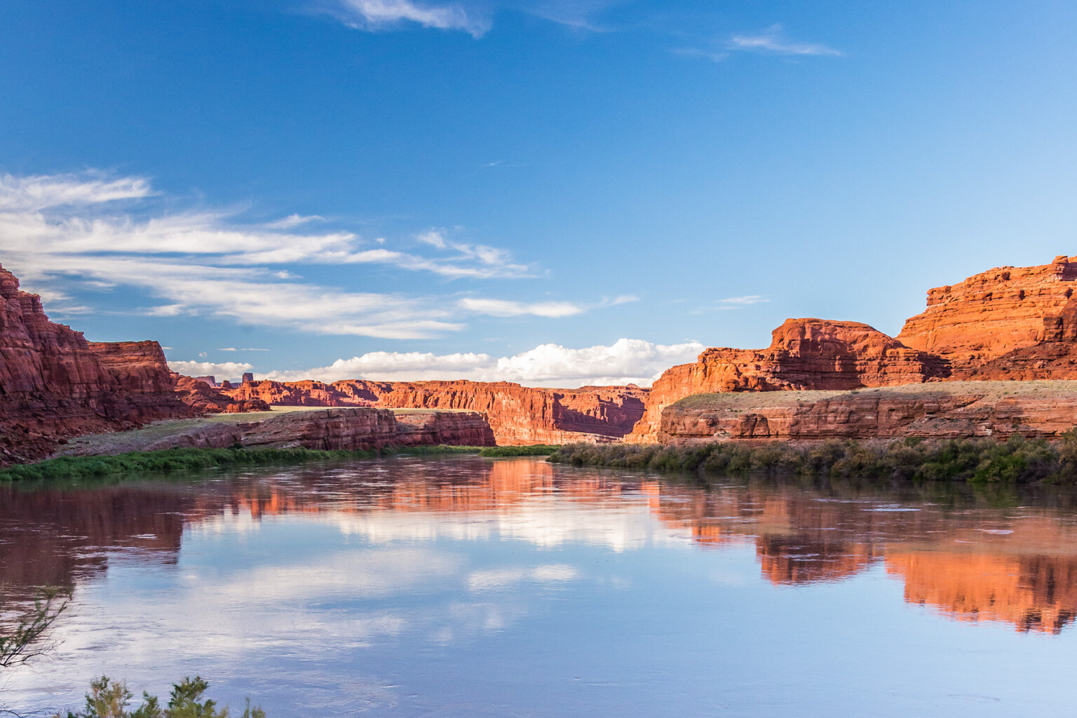 Vast redstone canyon walls surround the Colorado River and are reflected in the water.