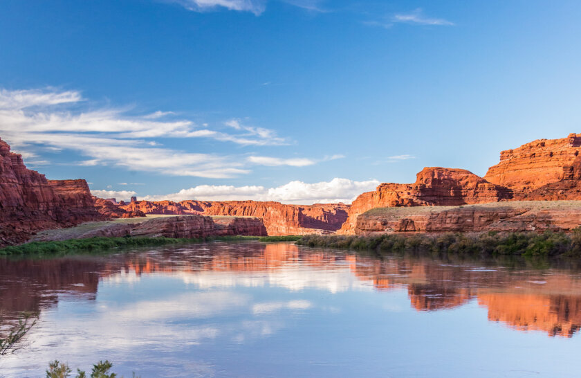 Vast redstone canyon walls surround the Colorado River and are reflected in the water.