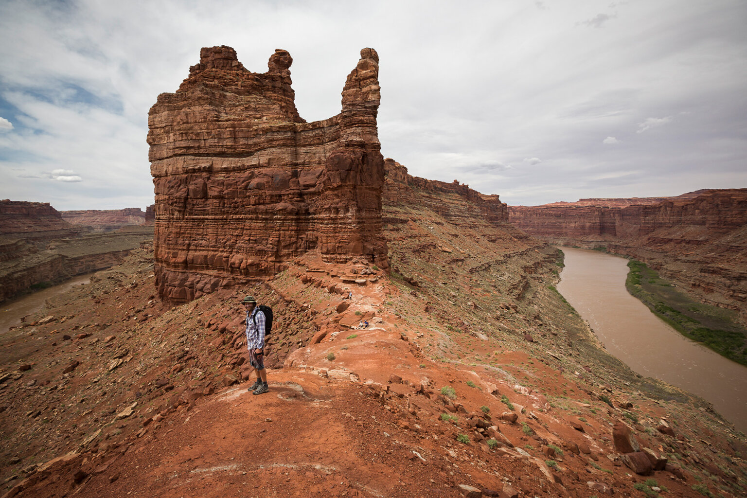 A hiker stands high up on a hill in front of sandstone canyon walls rising up and the river can be seen on both sides.