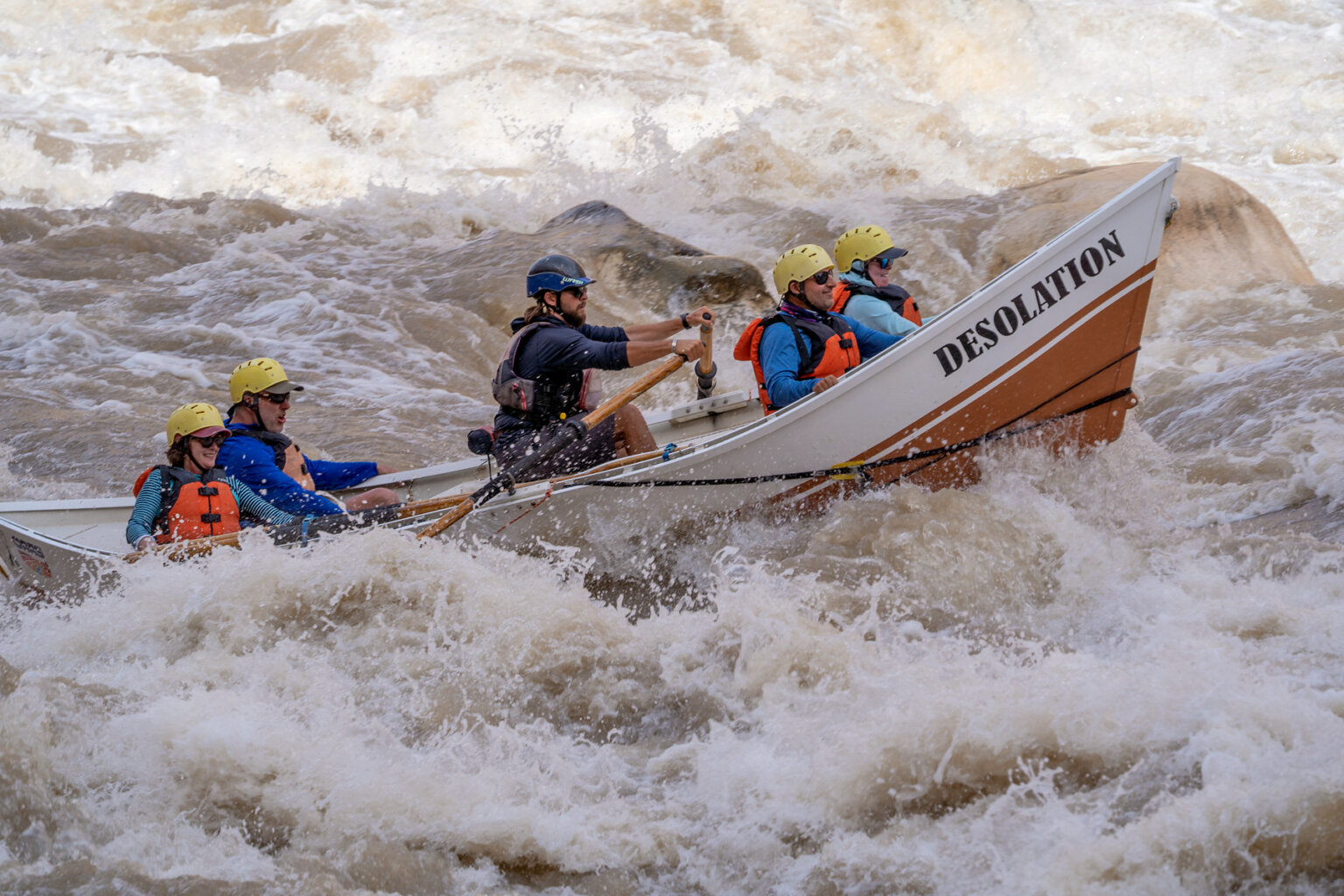 Side view of a dory rowed by a guide with four rafters holding on as they crash through a wave in a rapid on the Colorado River in Cataract Canyon.