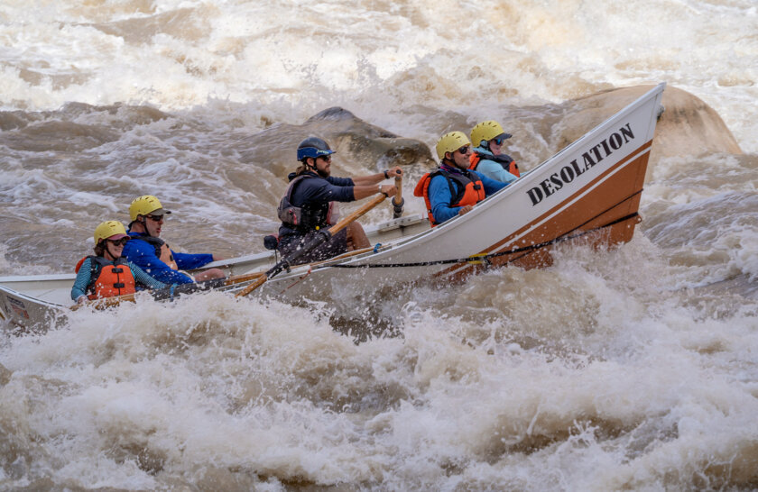 Side view of a dory rowed by a guide with four rafters holding on as they crash through a wave in a rapid on the Colorado River in Cataract Canyon.