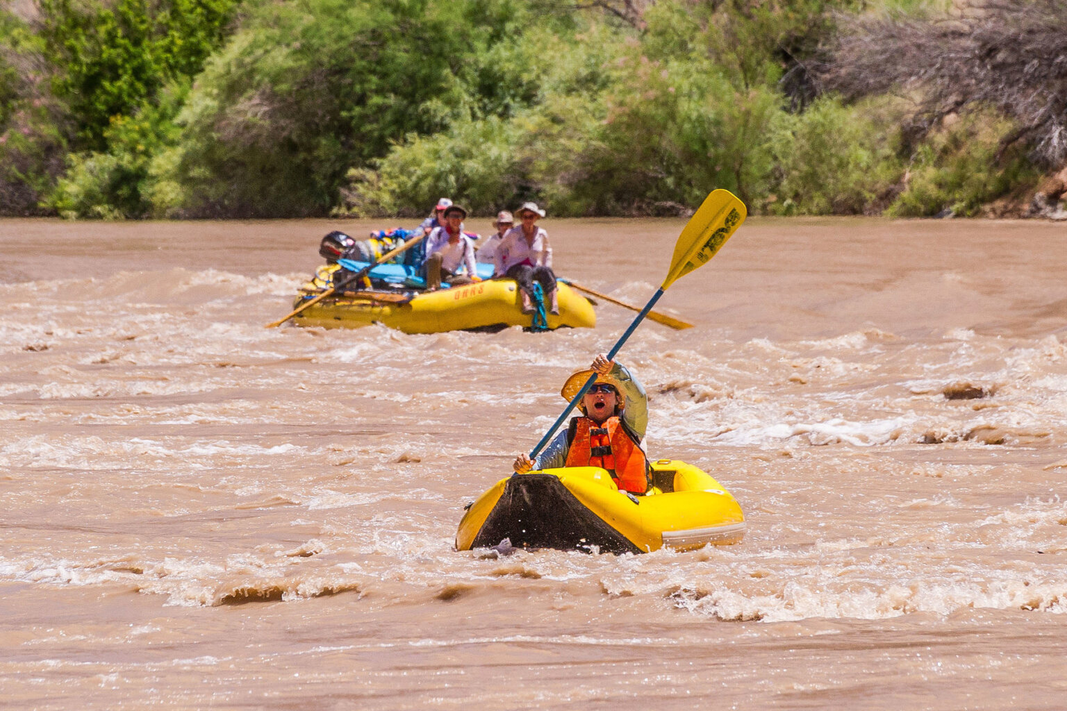 An excited kayaker paddles on the Colorado River while a raft of guests behind them watch.