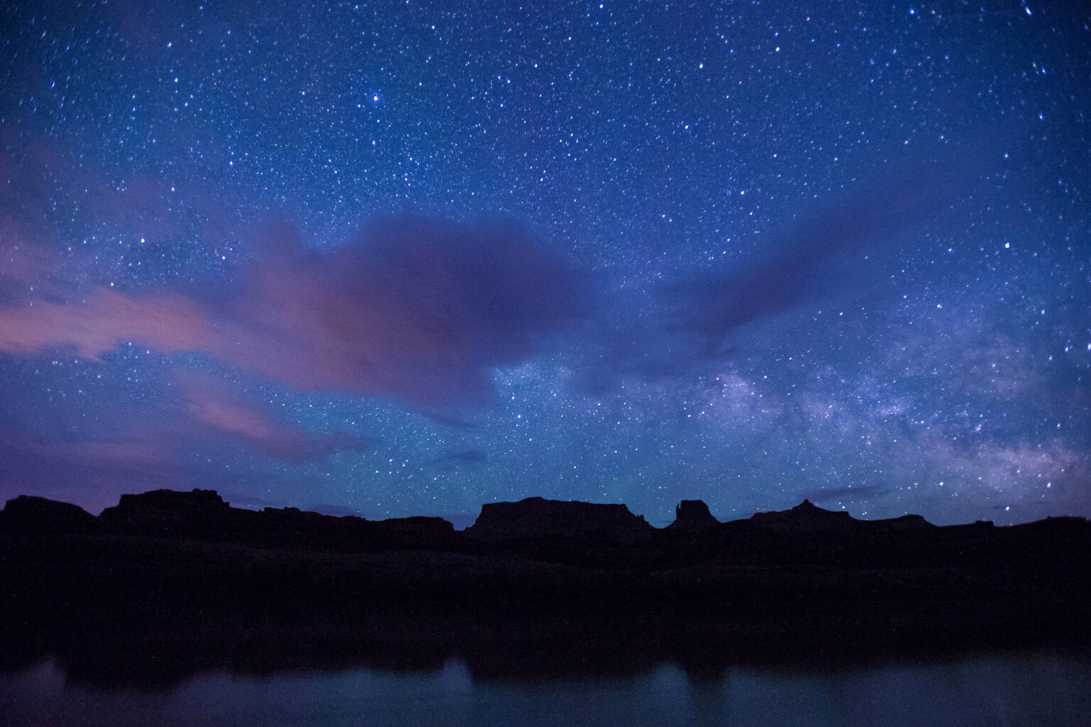 A vast starry sky with whispy clouds above the black silhouette of canyon walls across the river.