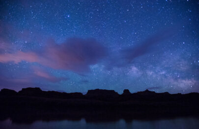 A vast starry sky with whispy clouds above the black silhouette of canyon walls across the river.