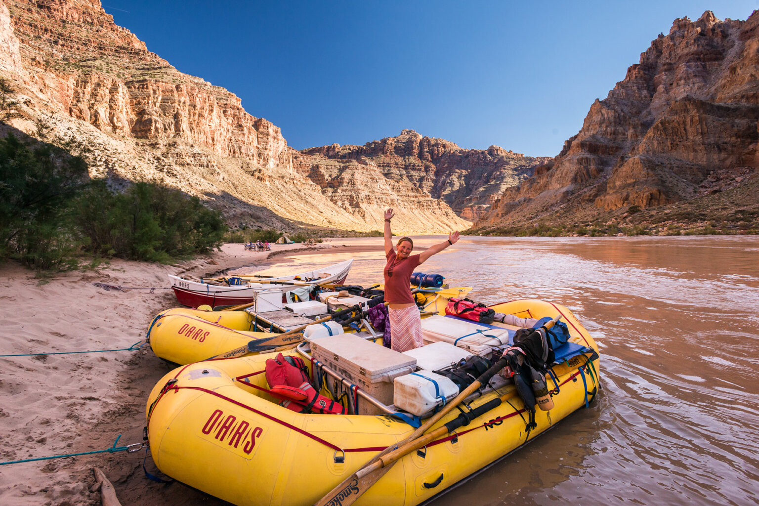 A guide stands on her raft and smiles at the camera with her hands in the air along the banks of the Colorado River.