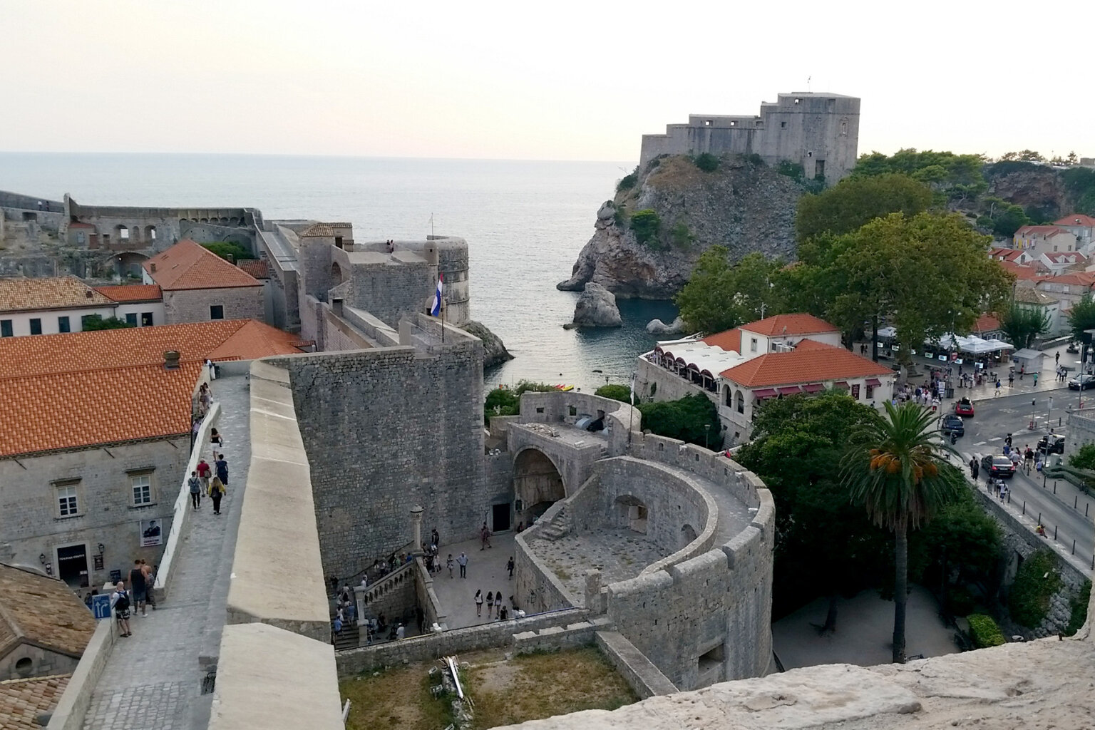 View of one of the charming sea-side town in Croatia as people walk along it's streets with a castle perched high on a hill in the background.