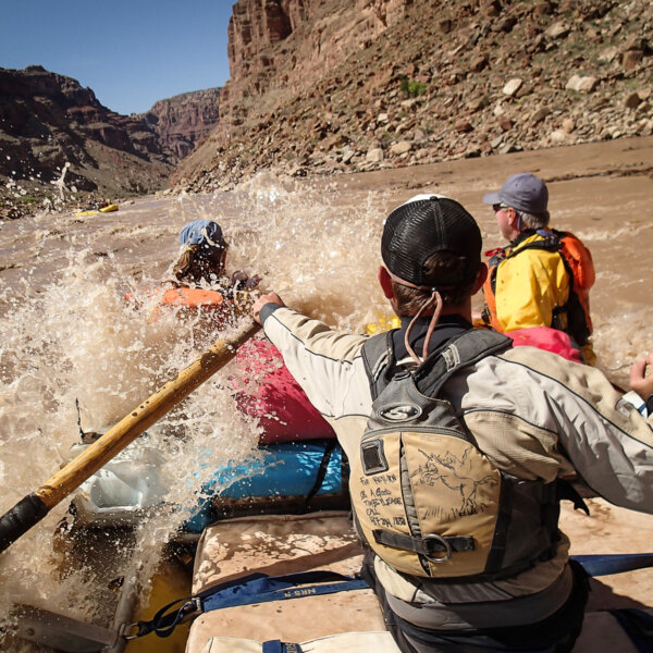 A guide rows an oar raft through a rapid on the Colorado River as passengers in the front are splashed by the waves.