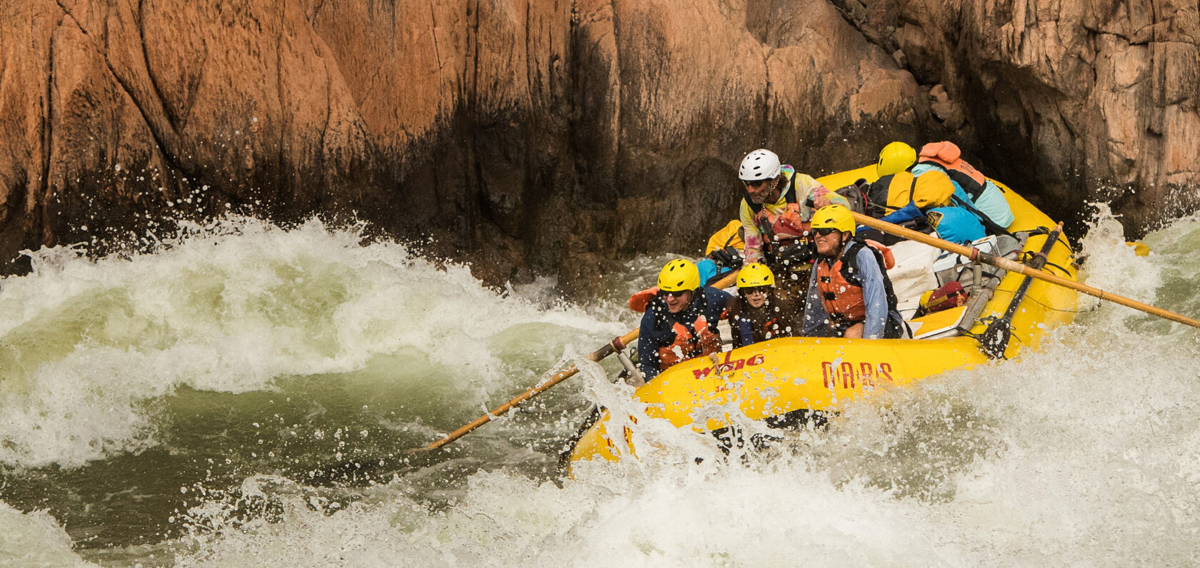 A man pilots a raft and his passengers through a large, splashy rapid on the Colorado River in Grand Canyon