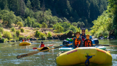 Two kids sit on the front of a yellow raft as they float on a calm stretch of the Rogue River