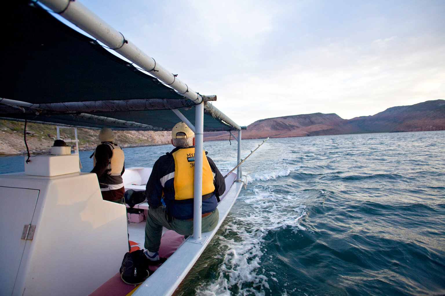 A motor boat on the ocean and a passenger is sitting on the edge of the boat fishing.