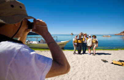 A group of guests stand on the beach with the ocean behind them as another guests takes their picture.