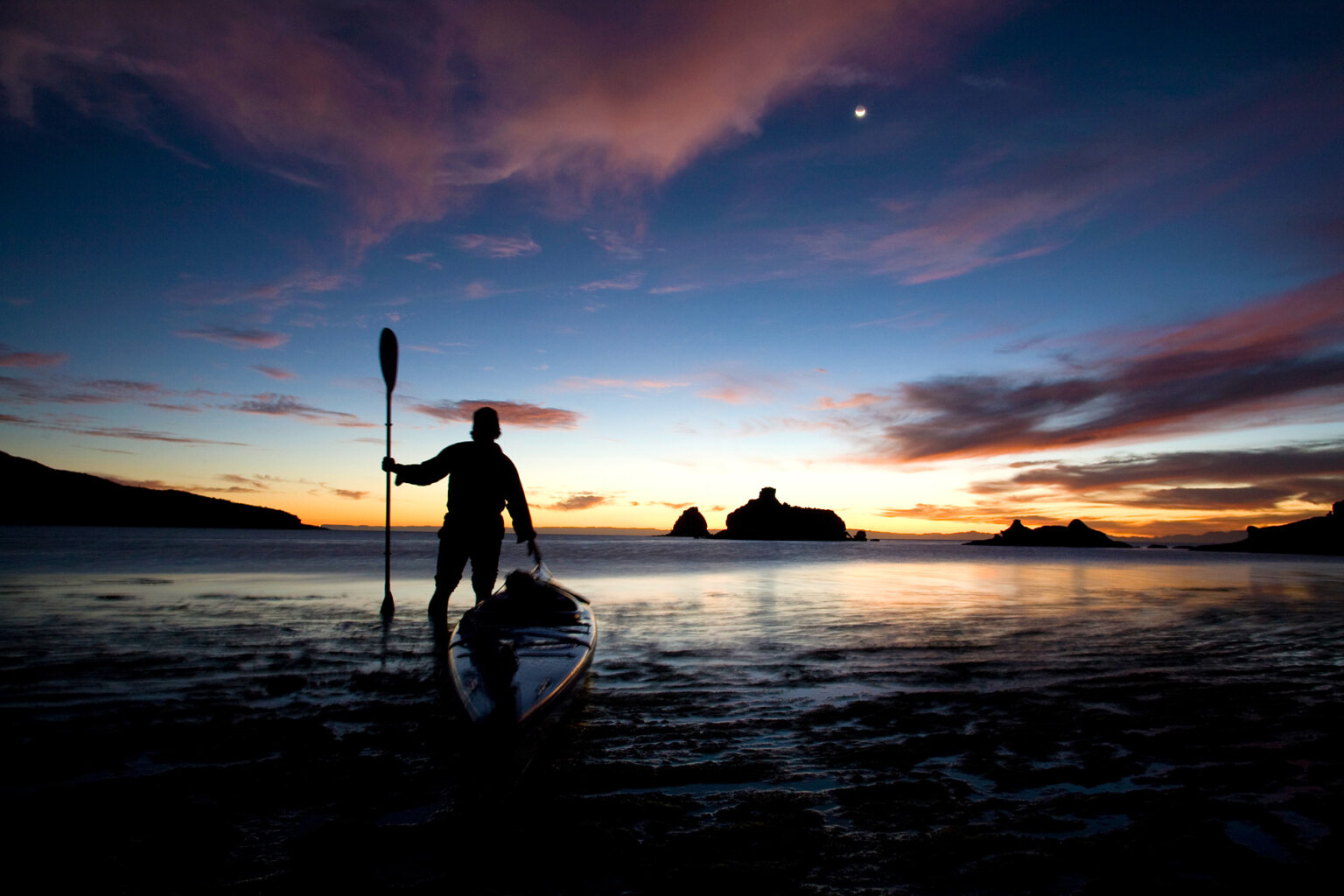 Silhouette of a person next to a sea kayak with their paddle held up straight in their hand as the sun sets on the ocean behind them.