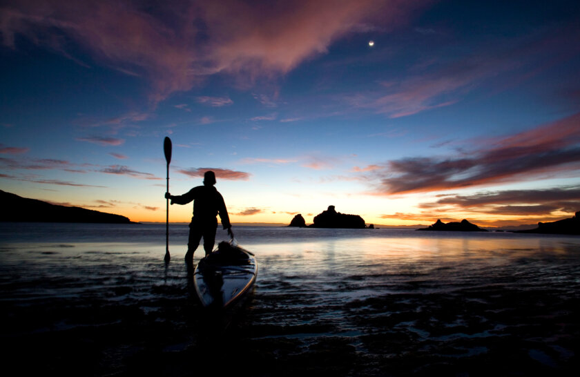 Silhouette of a person next to a sea kayak with their paddle held up straight in their hand as the sun sets on the ocean behind them.