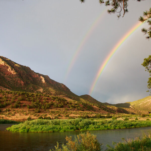A double rainbow is seen above a riverscape with mountains studded with lots of green vegetation.