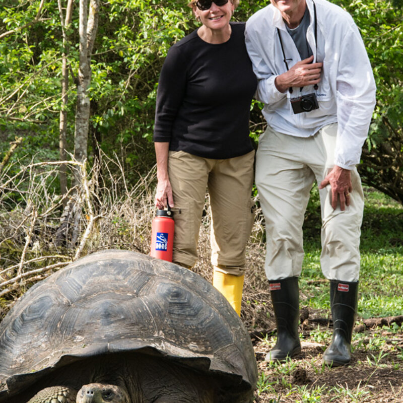 A couple stands behind a giant tortoise and smiles for the camera in the Galapagos Islands.