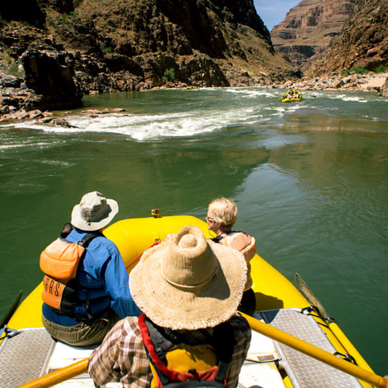View from behind of a guide rowing with two guests sitting in front in a yellow raft as they head downstream on the Colorado River through Grand Canyon.