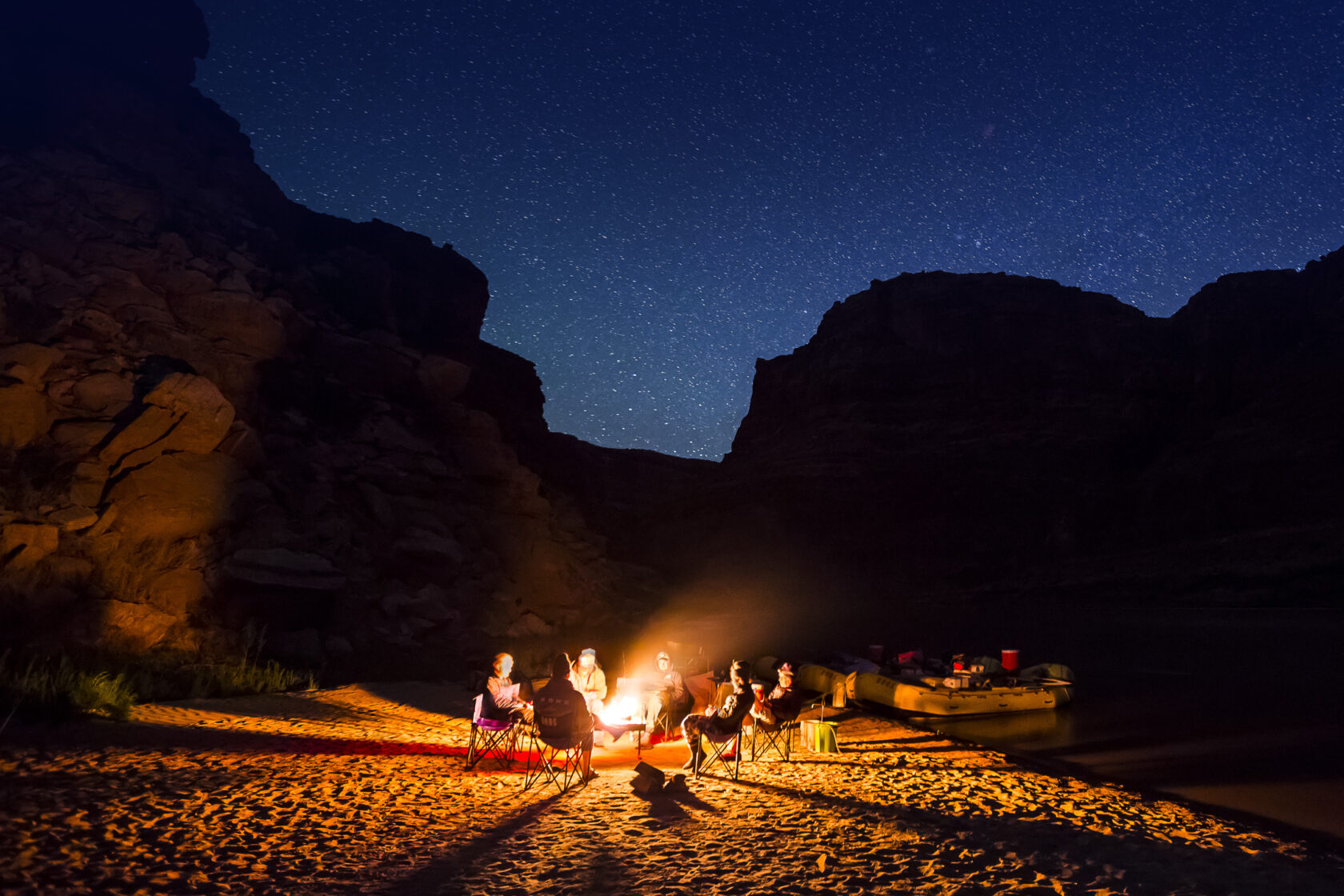 A group of friends sits around a campfire at night, surrounded by rocky cliffs under a starry sky, with rafts nearby. Canyonlands National Park.