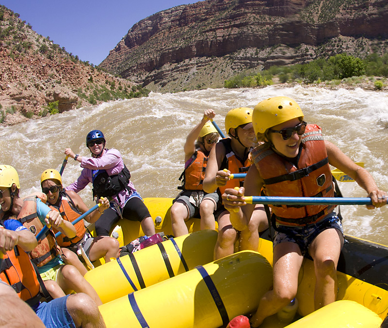 A group of people in yellow helmets paddles vigorously in a bright yellow raft, navigating a turbulent river surrounded by rocky cliffs.
