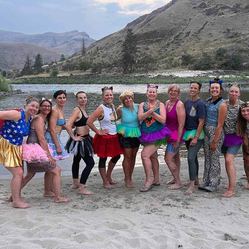 A group of smiling women in costumes lined up on a sandy beach on an OARS Gorges of the Lower Salmon River Women's Wellness trip.