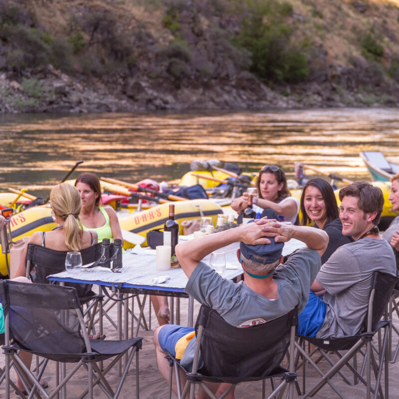 A group of adults sit around a linen draped table on a sandy beach drinking wine and socializing on an OARS Wine on the Salmon River trip.