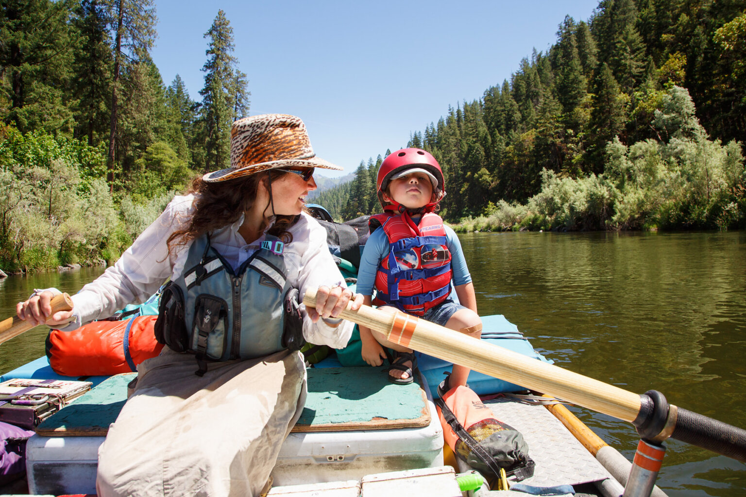 A female guide rowing downstreamon the Lower Klamath River, looking over her shoulder and smiling at a young child sitting just behind her.
