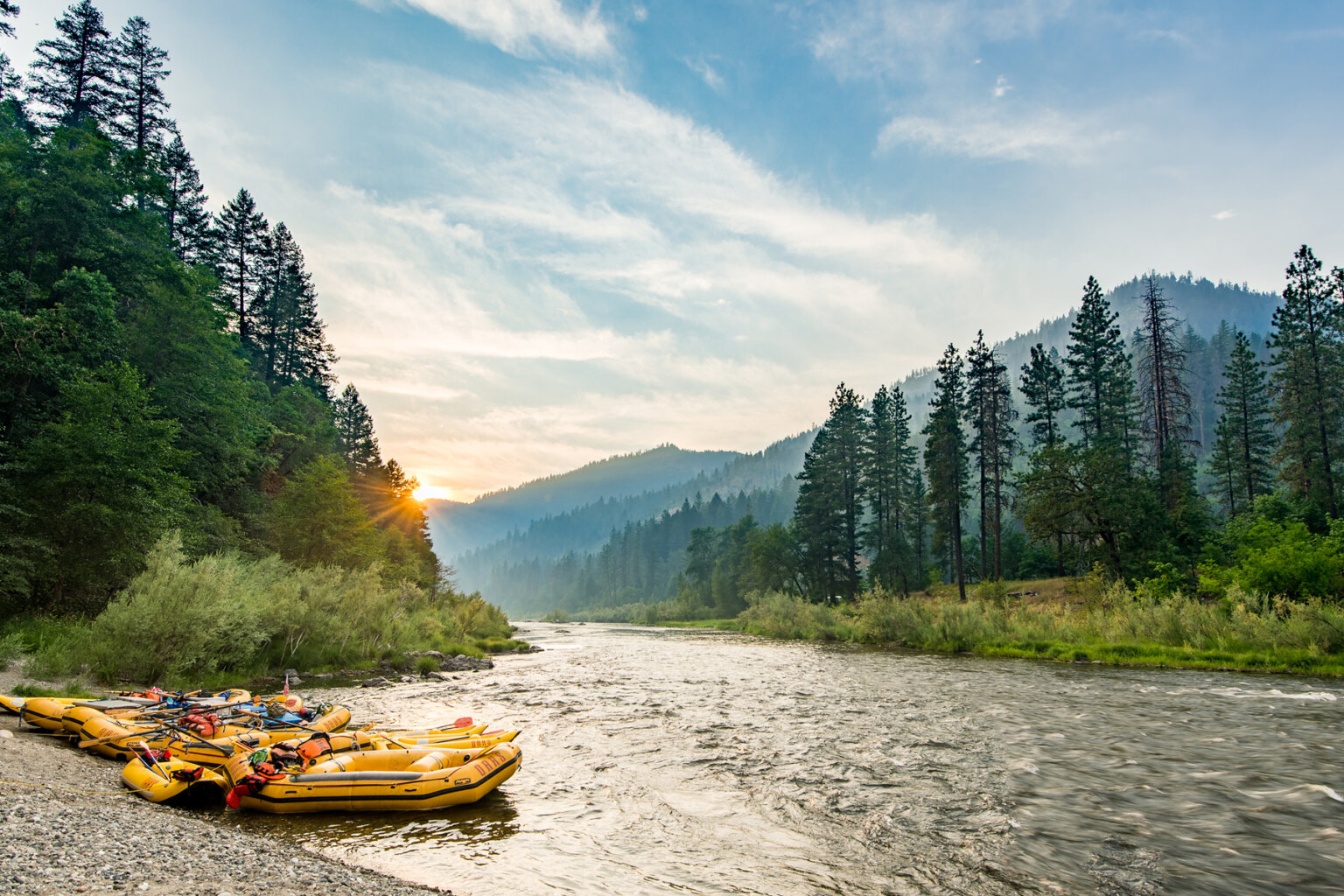 Several yellow rafts tied up at the shoreline at sunset with verdant forested hills and the Lower Klamath River as the backdrop.
