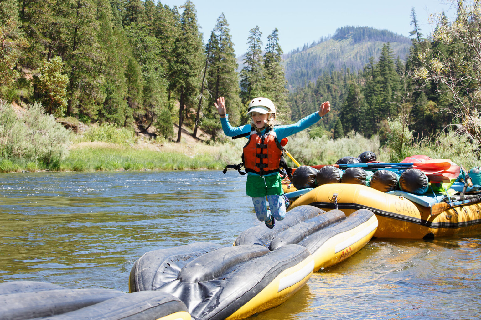 Three inflatable kayaks are turned upside down and lashed together front to back to a large oar raft as an obstacle course game; a young girl is jumping mid-air off of it into the river.
