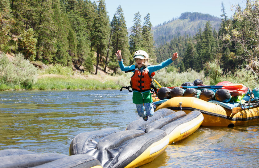 Three inflatable kayaks are turned upside down and lashed together front to back to a large oar raft as an obstacle course game; a young girl is jumping mid-air off of it into the river.