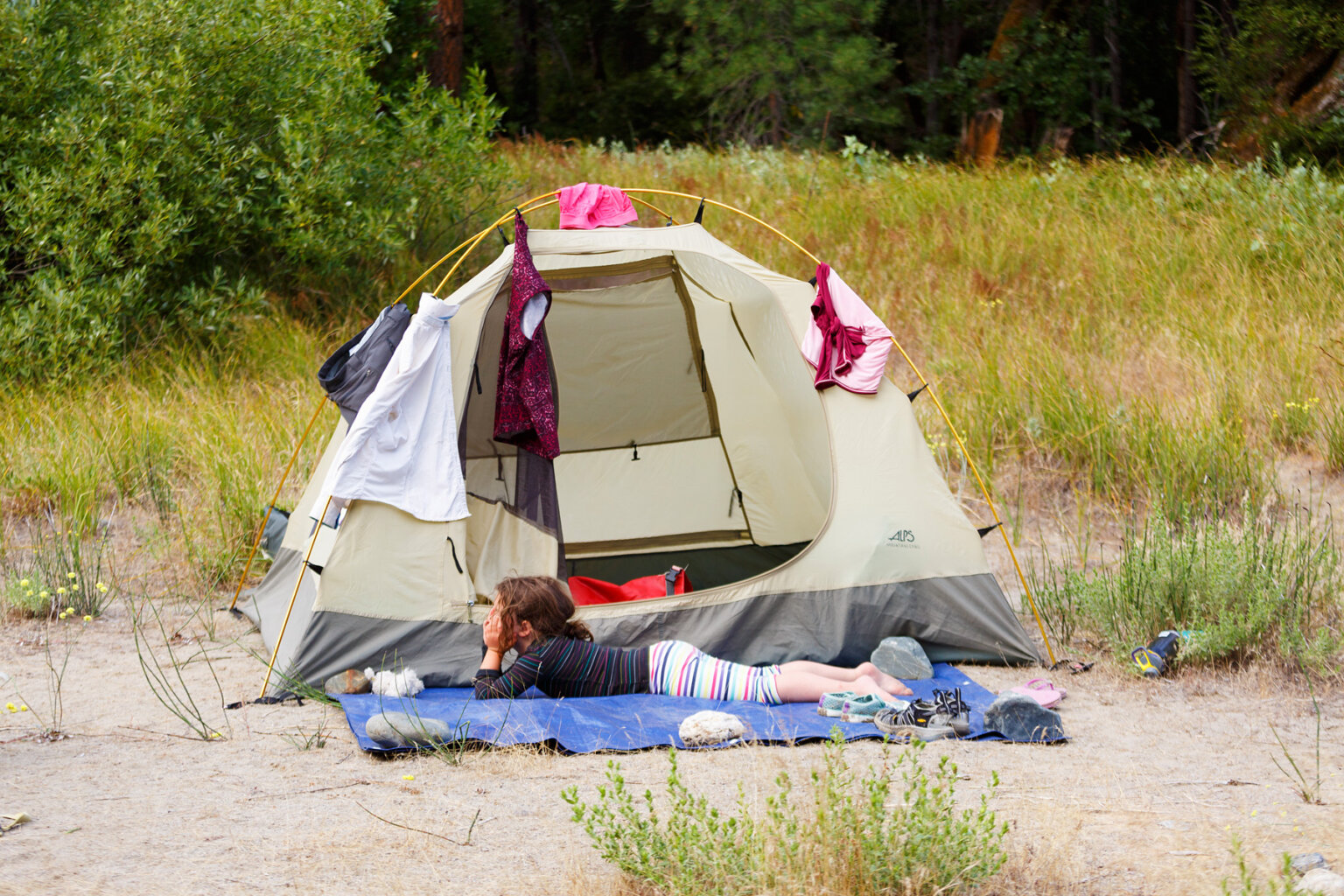 A young barefoot girl lays in front of her tent on a ground tarp with her head in her hands looking at the scenery; serveral articles of clothing are drying on the tent.