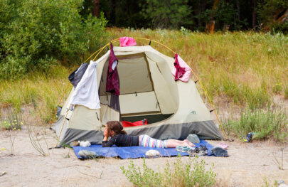 A young barefoot girl lays in front of her tent on a ground tarp with her head in her hands looking at the scenery; serveral articles of clothing are drying on the tent.