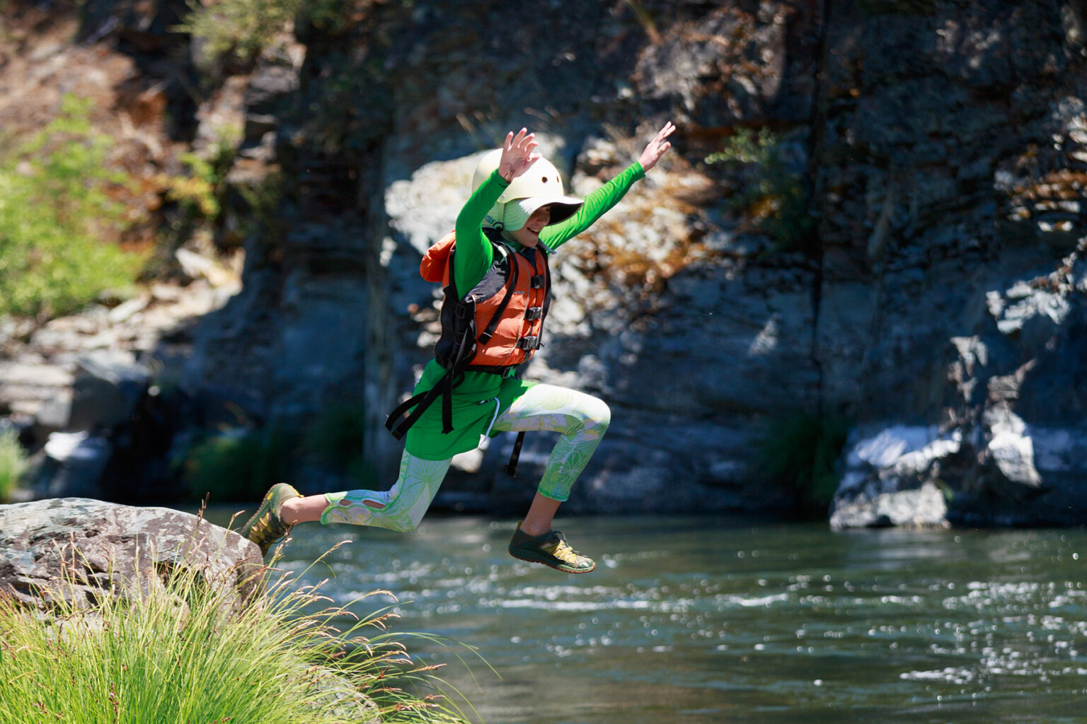 A kid jumps off a boulder into the river with their hands in the air above them and a smile on their face.