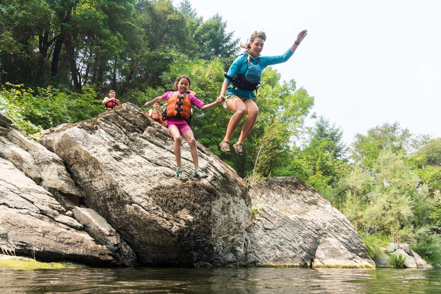A guide holds the hand of a smiling young girl and are mid-air having just jumped off a large boulder into the river.