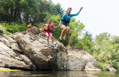 A guide holds the hand of a smiling young girl and are mid-air having just jumped off a large boulder into the river.