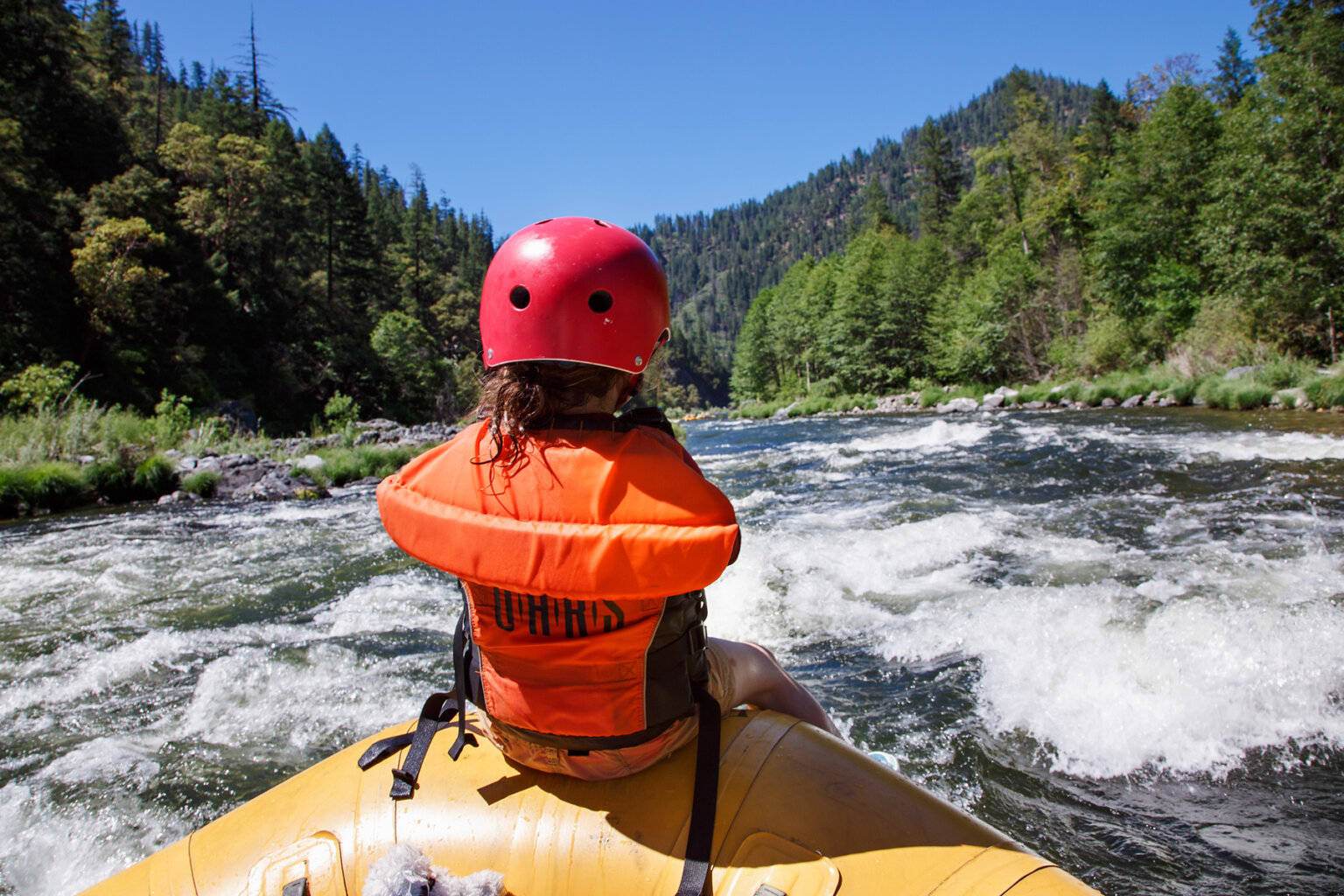 View from behind of a kid wearing an orange PFD and red helmet riding the front of a raft as it enters a stretch of small rapids.