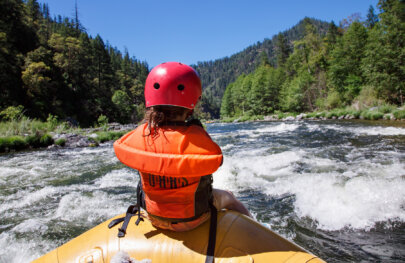 View from behind of a kid wearing an orange PFD and red helmet riding the front of a raft as it enters a stretch of small rapids.