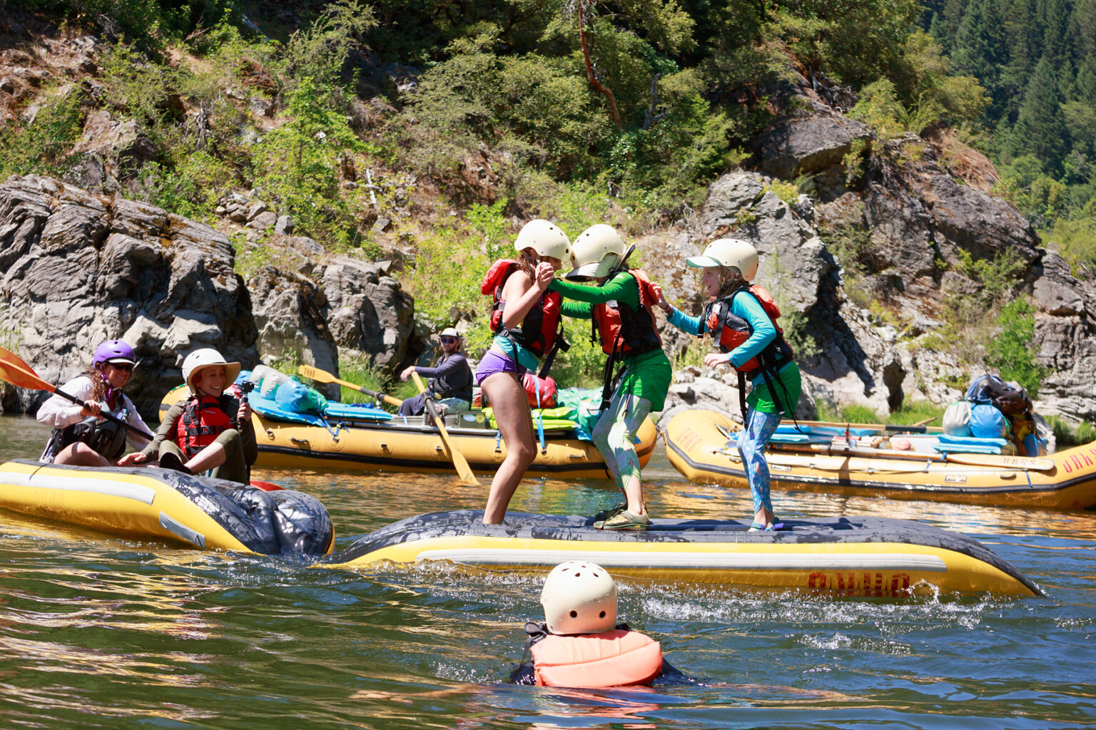 Two inflatable kayaks are turned upside down in the river and lashed together front to end; on one sits two adults as they watch three children standing on the other trying to keep their balance as an obstacle course river game.