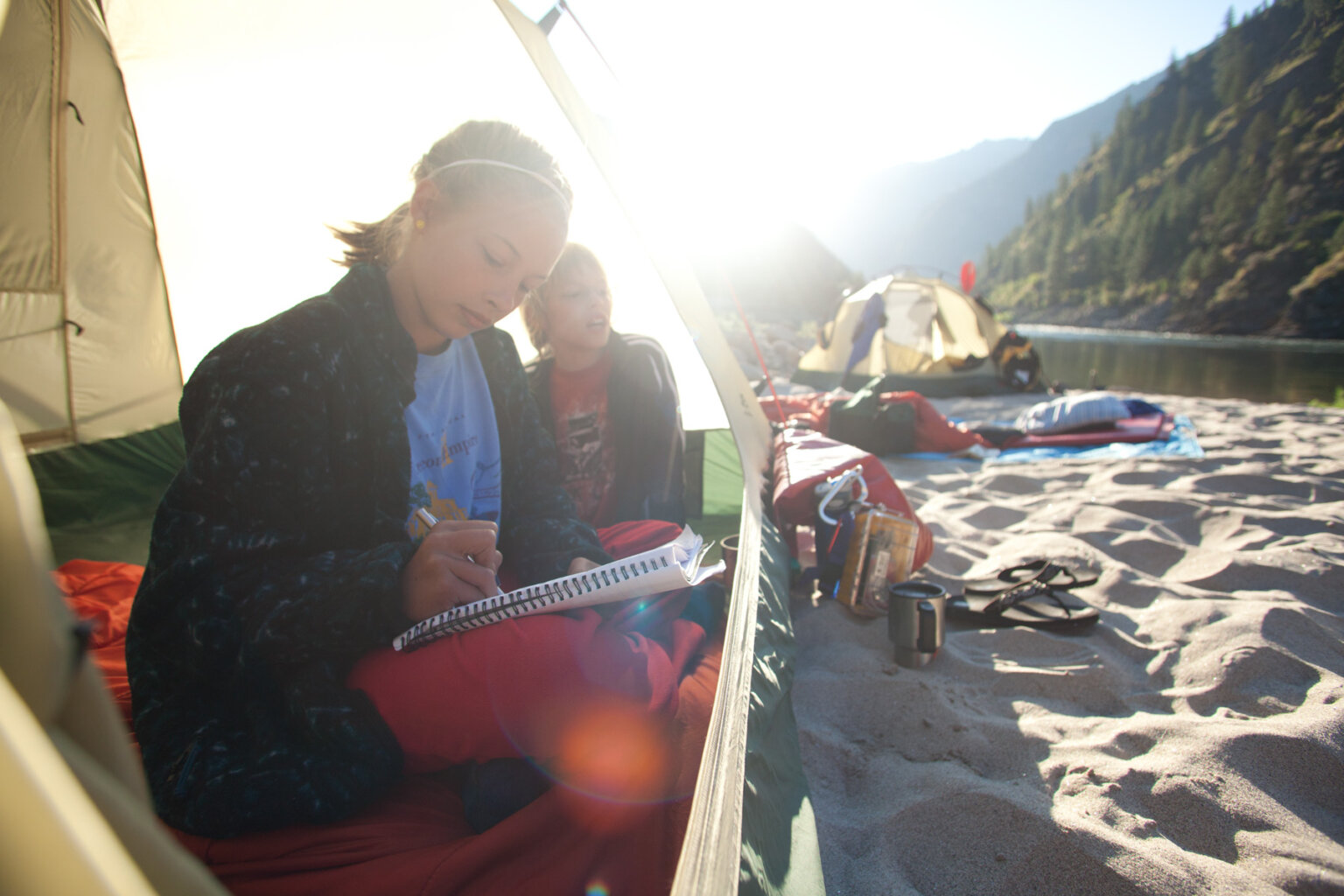 A young woman and young boy sit just inside a tent while the young woman writes in a notebook and the setting sun behind them shines through the tent fabric.