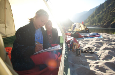 A young woman and young boy sit just inside a tent while the young woman writes in a notebook and the setting sun behind them shines through the tent fabric.