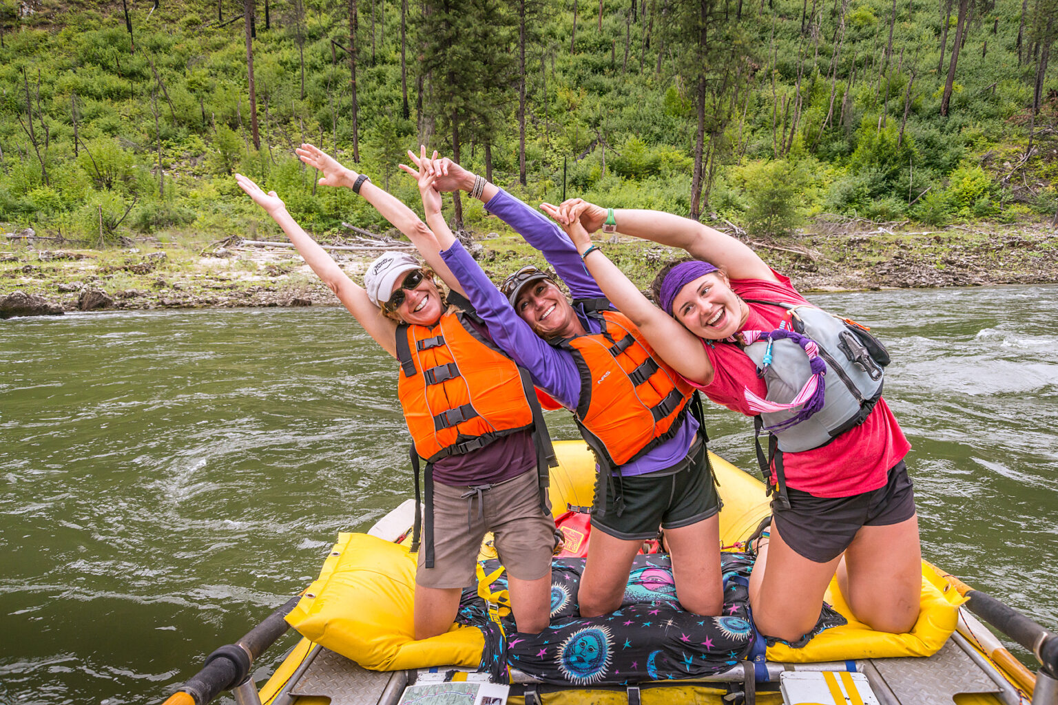 Three women on their knees lined up on sleeping pad strapped to a raft all leaning the same way as they smile and strike a yoga pose.