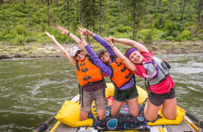 Three women on their knees lined up on sleeping pad strapped to a raft all leaning the same way as they smile and strike a yoga pose.