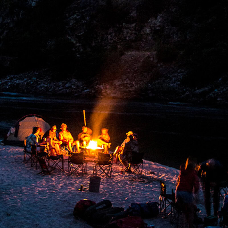 A night shot of a group of people sitting around a small fire in a fire pan on a sandy beach at camp on the Main Salmon River.