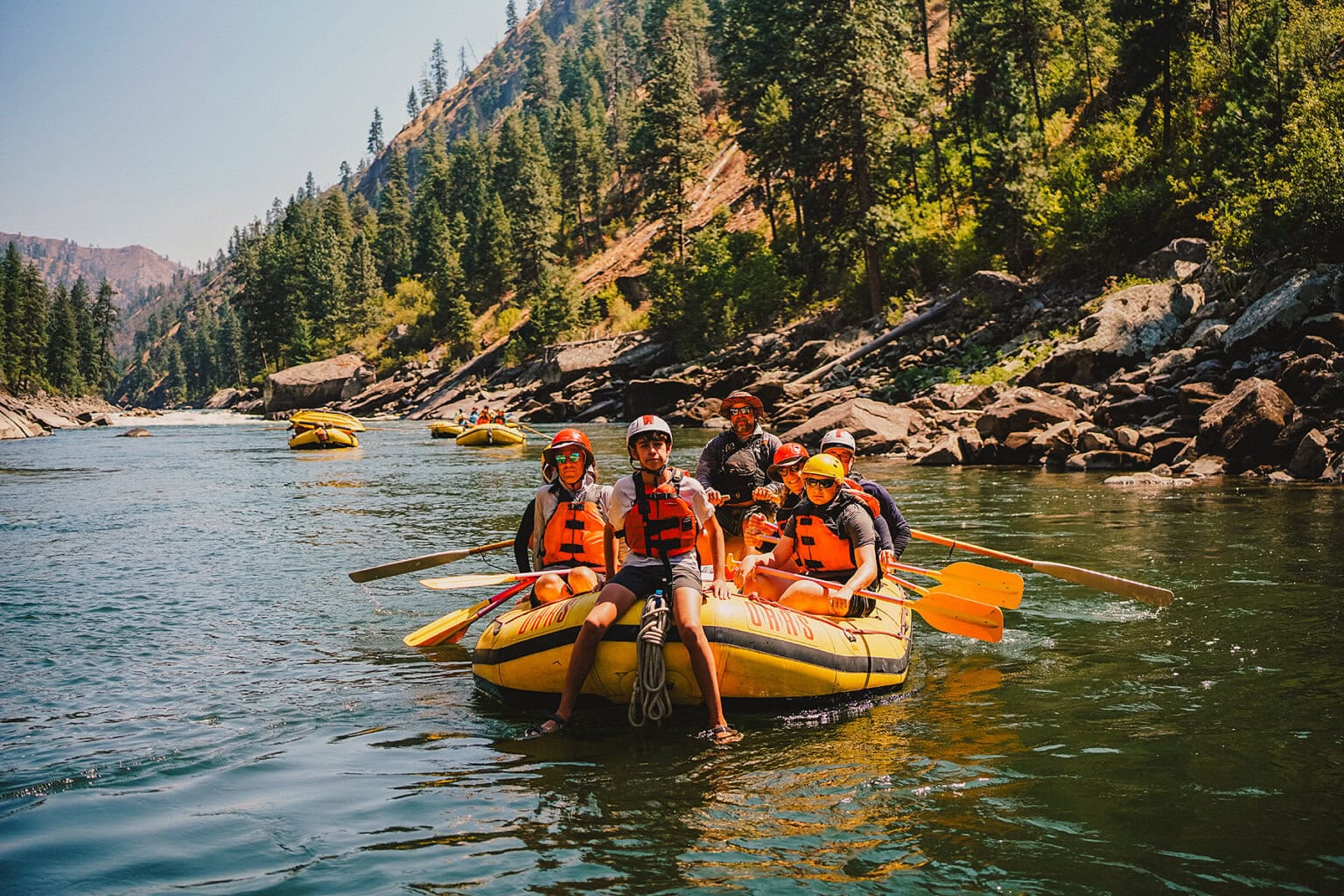 A group of six people, wearing life jackets and helmets, enjoy a river rafting adventure on the Main Salmon River on a sunny day, surrounded by lush greenery.