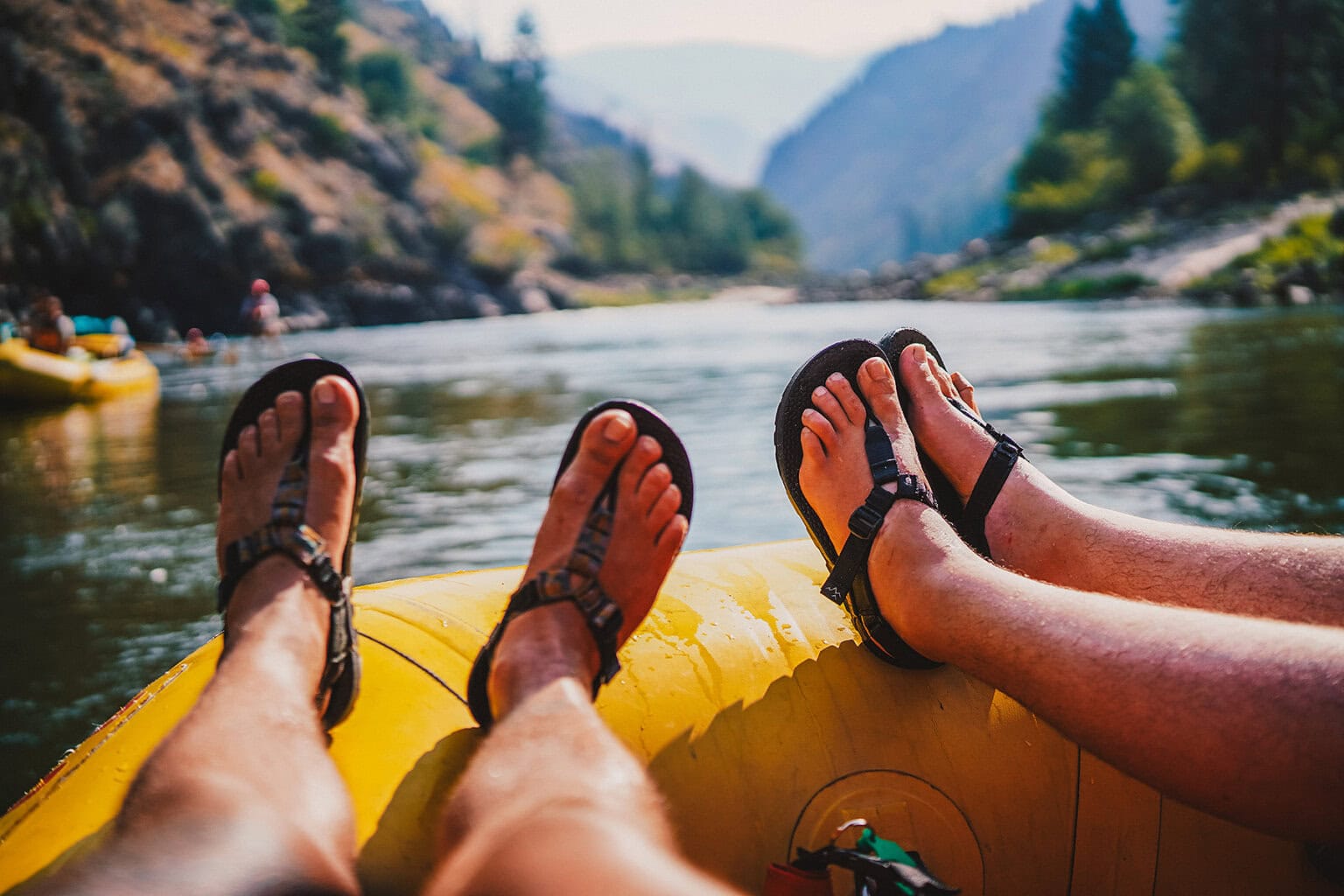 A close-up of two pairs of feet in sandals relaxed on the edge of a yellow raft, floating down a serene river surrounded by mountains.