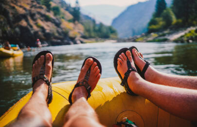 A close-up of two pairs of feet in sandals relaxed on the edge of a yellow raft, floating down a serene river surrounded by mountains.